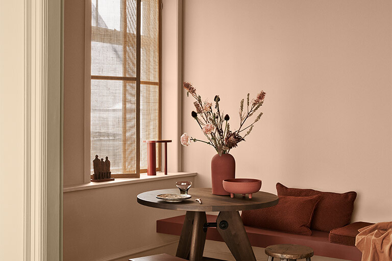 Ljust rum med rosa väggar och ett litet träbord med bukett i vas, bredvid pallar och bänk