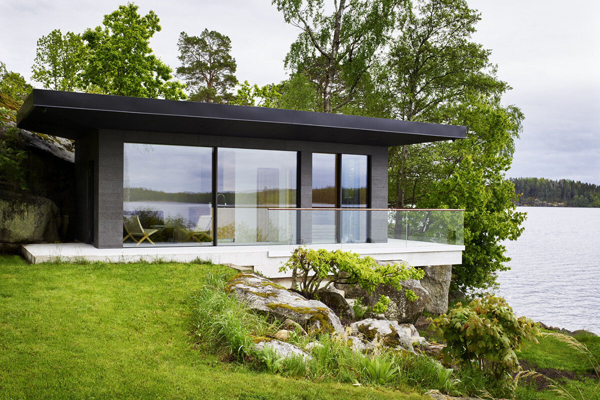 Ett svart mindre uthus på en klippkant utsikt över vatten, skog och grön gräsmatta